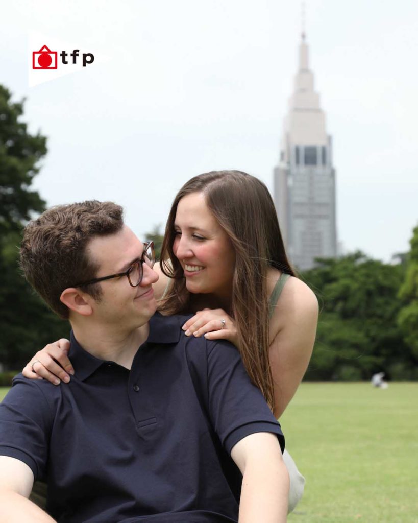 Tokyo’s Green Parks2 alt="Engagement photo of a couple on the grass in central Tokyo with a city tower in the background, smiling and embracing each other"